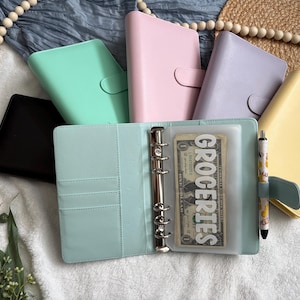 May include: A collection of six A6-sized ring binders in black, mint green, pastel pink, lavender, and yellow. One open mint green binder features a metal six-ring mechanism, a pen holder with a patterned pen, and a clear plastic pouch labeled GROCERIES.