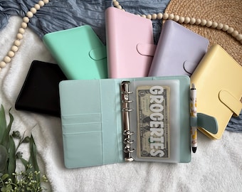 May include: A collection of six A6-sized ring binders in black, mint green, pastel pink, lavender, and yellow. One open mint green binder features a metal six-ring mechanism, a pen holder with a patterned pen, and a clear plastic pouch labeled GROCERIES.