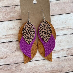 May include: A pair of dangle earrings with three layers of leather. The top layer is a leopard print, the middle layer is a solid purple, and the bottom layer is a brown textured leather.