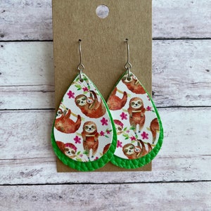 May include: Teardrop-shaped earrings featuring a white background with a repeating pattern of sloths and pink flowers. The earrings have a green border and silver hooks. The earrings are displayed on a brown card.