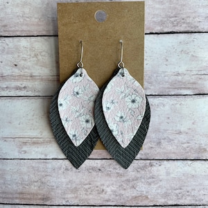 May include: A pair of dangle earrings with a layered design. The top layer is pink with a white floral pattern, and the bottom layer is gray. The earrings are made of leather and have silver hooks.