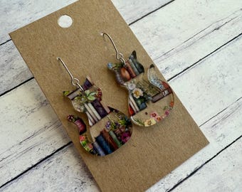 May include: A pair of cat-shaped earrings with a bookshelf design. The earrings are made of clear acrylic and feature a colourful image of books, flowers, and other objects. The earrings are hanging on a brown card.