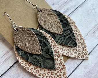 May include: A pair of dangle earrings with three layers of faux leather. The top layer is a gold glitter leaf shape, the middle layer is a gray and black snake skin pattern, and the bottom layer is a white and brown leopard print.