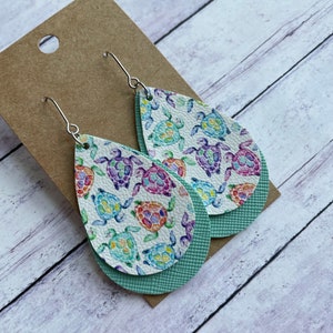 May include: Teardrop-shaped earrings featuring a white background with colorful sea turtles in shades of blue, purple, and orange. The earrings have a textured, light green backing and silver-colored ear wires. The earrings are displayed on a brown card.