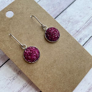 May include: A pair of dangle earrings featuring round, magenta-colored druzy stones set in silver-toned metal. The earrings are displayed on a brown earring card against a light wood-grain background.