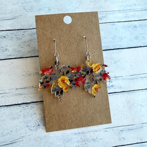 May include: A pair of dangle earrings featuring clear acrylic maple leaf shapes with a printed floral pattern. The design includes yellow roses, red flowers, and dark blue foliage. The earrings hang from silver-toned metal hooks on a brown card.