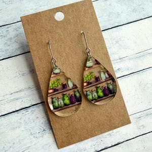 May include: Clear, teardrop-shaped earrings with a silver hook. The earrings feature a colorful illustration of shelves filled with jars and plants. The jars are green and purple. The earrings are displayed on a brown card.