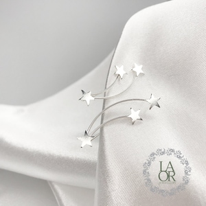 Puede incluir: Pendientes de plata en forma de estrella fugaz con cinco colgantes de estrella. Los pendientes están sobre un fondo de tela blanca. LA OR JEWELLERY está impreso en un logotipo circular en la esquina inferior derecha.