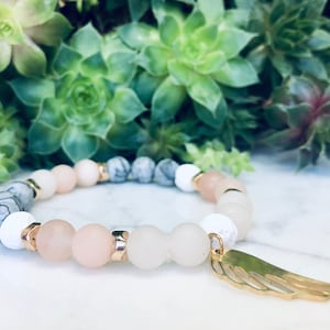 Peut inclure: Un bracelet délicat avec des perles blanches, grises et roses et un pendentif en forme d'aile d'ange dorée. Le bracelet est fait de pierres naturelles et est parfait pour tous les jours.