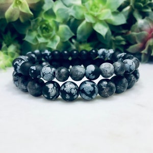 May include: Two black and white snowflake obsidian beaded bracelets. The bracelets are stacked on top of each other.