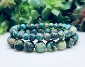 May include: Two turquoise gemstone bracelets with a speckled pattern. The bracelets are made of round beads and are stacked on top of each other.