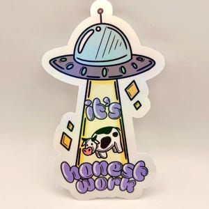 May include: A sticker depicting a light blue UFO with a cow being abducted. The text "it's honest work" is written in purple script below the UFO and cow graphic. The sticker has a white border and a small hole at the top.