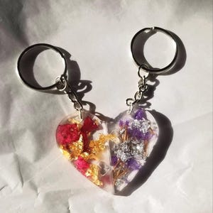 May include: A pair of heart-shaped keychains with a puzzle piece design. One half features red and gold pressed flowers, while the other half features purple and silver pressed flowers.