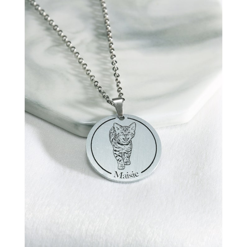 Pu&ograve; includere: Collana d'argento con un ciondolo rotondo che presenta un'immagine incisa in bianco e nero di un gatto. Il ciondolo ha il nome "Maisie" inciso sotto l'immagine.