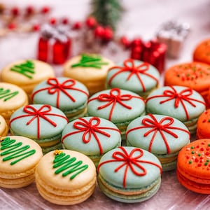 May include: A collection of festive macarons in various colors. Some are light green with red bow designs, others are orange with green and red sprinkles, and some are yellow with green Christmas tree designs.
