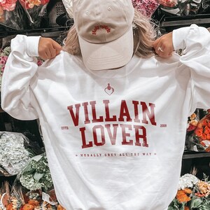 May include: White sweatshirt with red text that reads "VILLAIN LOVER" with the years 1978 and 2008 on either side of the text. The text also includes "MORALLY GREY ALL THE WAY" in smaller font. A person is wearing the sweatshirt with a beige baseball cap that says "Holy Water" on it.
