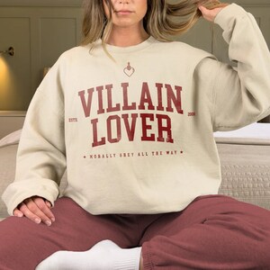 May include: A cream-coloured sweatshirt with a red graphic that says "VILLAIN LOVER" with the text "ESTD. 2008" and "MORALLY GREY ALL THE WAY" in smaller font.