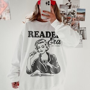May include: White sweatshirt with a black and white illustration of a woman reading a book and drinking from a glass. The text "Reader Era" is printed above the illustration and the text "I can't, I'm booked" is printed below the illustration.