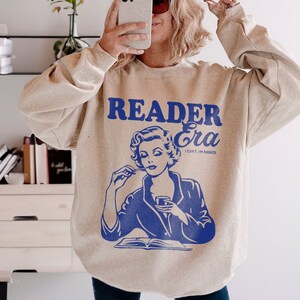 May include: A beige sweatshirt with a blue graphic of a woman reading a book and drinking from a glass. The text on the sweatshirt reads "Reader Era I can't I'm booked".