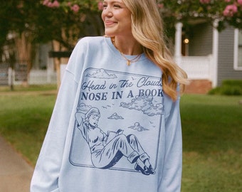 May include: A light blue sweatshirt with a graphic print of a woman reading a book in the clouds. The text on the sweatshirt reads "Head in the Clouds Nose in a Book."