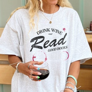Op de afbeelding: Een grijs T-shirt met de tekst "Drink Wine and Read Good Books" in zwarte letters. Het shirt heeft een verweerde look en rode wijnvlekken.
