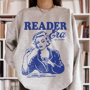 May include: Gray sweatshirt with a blue graphic of a woman reading a book and the text "Reader Era I Can't, I'm Booked".