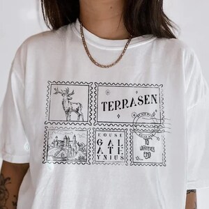 May include: White oversized t-shirt with a graphic print of four postage stamps. The stamps feature a deer, a castle, the words "Fouse Gal Athis Ynius", and the words "Terrasen By Air Mail" and "To Whatever End".