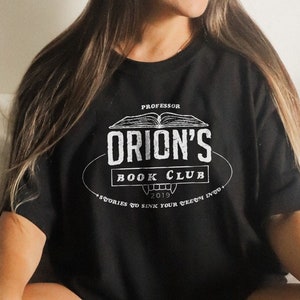 K&ouml;nnte beinhalten: Schwarzes T-Shirt mit dem wei&szlig;en Text "Professor Orion's Book Club 2019". Das Design zeigt ein offenes Buch. Die Person tr&auml;gt das T-Shirt und sitzt auf einem Bett mit einem Buch und einem Weidenkorb.