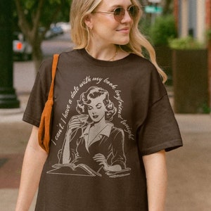 May include: A brown t-shirt with a vintage illustration of a woman reading a book and drinking from a cup. The text on the shirt reads "I can't I have a date with my book boyfriend tonight."