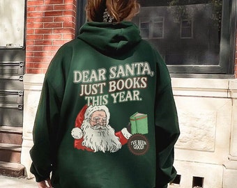 May include: A person wearing a green hooded sweatshirt with the text "Dear Santa, Just Books This Year." and a cartoon image of Santa Claus. The sweatshirt is being worn with light blue jeans and white sneakers.