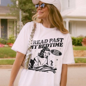 Op de afbeelding: Wit T-shirt met een zwart-witte illustratie van een vrouw die een boek leest. De tekst op het shirt luidt "Read Past My Bedtime" en "Midnight Readers Book Club".