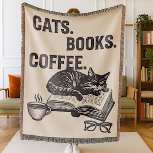 May include: Beige throw blanket with black text that reads "CATS. BOOKS. COFFEE." A black and white illustration of a cat sleeping on a book, a coffee cup, and glasses are also featured. The blanket has a fringed edge.