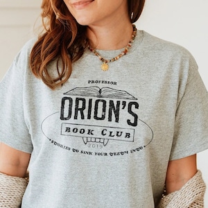K&ouml;nnte beinhalten: Ein meliertes graues T-Shirt mit dem schwarzen Schriftzug "Professor Orion's Book Club" und einer Abbildung eines offenen Buches. Das Shirt zeigt auch das Jahr "2019" und den Satz "Stories to sink your teeth into."