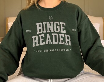 May include: A dark green sweatshirt with the text "Binge Reader" in white lettering. The sweatshirt has a book icon and the text "Estd. 2009" above the main text. The text "Just one more chapter" is printed below the main text.