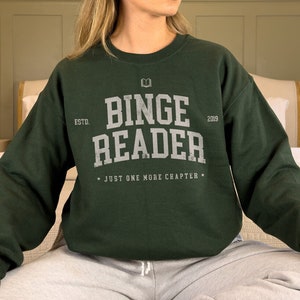 May include: A dark green sweatshirt with the text "Binge Reader" in white lettering. The sweatshirt has a book icon and the text "Estd. 2009" above the main text. The text "Just one more chapter" is printed below the main text.