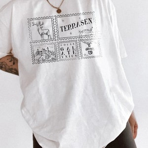May include: White oversized t-shirt with a graphic print of four postage stamps. The stamps feature a deer, a castle, the words "TERRASEN" and "GOUSE GAL ATHE YNIUS", and the words "TO WHATEVER END".