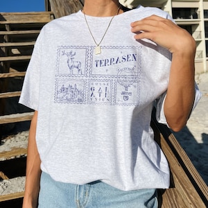 May include: Gray t-shirt with a blue graphic print of four postage stamps. The stamps feature a deer, a castle, the text "House Galathynus", and the text "To Whatever End".