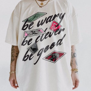Puede incluir: Camiseta blanca de gran tama&ntilde;o con un estampado gr&aacute;fico de cartas de juego y el texto "be wary be clever be good".