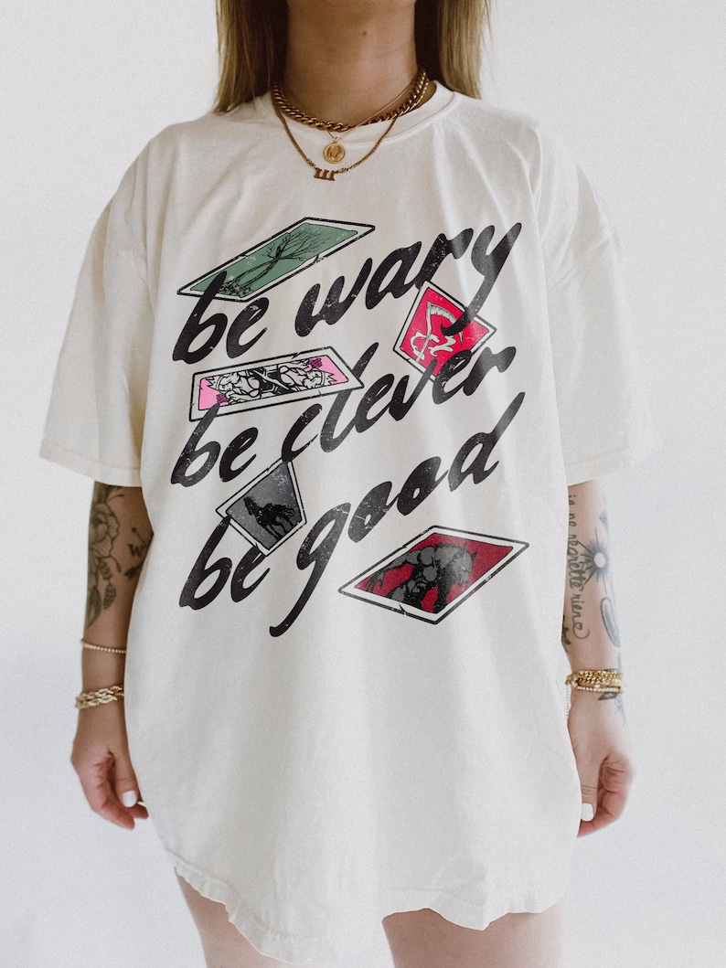 Puede incluir: Camiseta blanca de gran tama&ntilde;o con un estampado gr&aacute;fico de cartas de juego y el texto "be wary be clever be good".