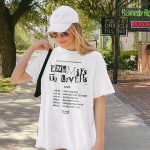 May include: White t-shirt with a black graphic that says "Enemies to Lovers" and lists dates and show titles. The dates are February 12, 14, 18, 21, 24, and 25. The show titles are "Only One Bed", "Who Did This to You?", "Fated Mates", "Slowest Burn", "Forced Proximity", and "I'll Kill for You".