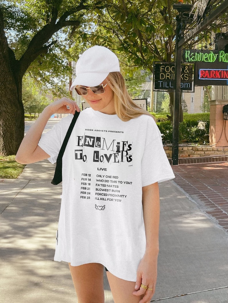 May include: White t-shirt with a black graphic that says "Enemies to Lovers" and lists dates and show titles. The dates are February 12, 14, 18, 21, 24, and 25. The show titles are "Only One Bed", "Who Did This to You?", "Fated Mates", "Slowest Burn", "Forced Proximity", and "I'll Kill for You".