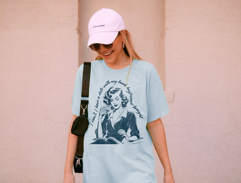May include: A light blue t-shirt with a vintage illustration of a woman reading a book. The text on the shirt reads "I can't I have a date with my book boyfriend tonight."