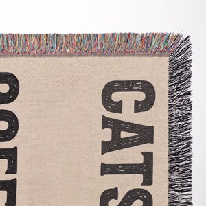 May include: A beige woven throw blanket with the words "COFFEE CATS" in large, black, block letters. The blanket has a fringed edge with a mix of colorful threads. The blanket is likely for home decor.