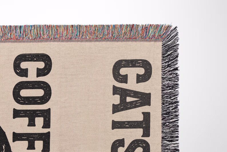 May include: A beige woven throw blanket with the words "COFFEE CATS" in large, black, block letters. The blanket has a fringed edge with a mix of colorful threads. The blanket is likely for home decor.