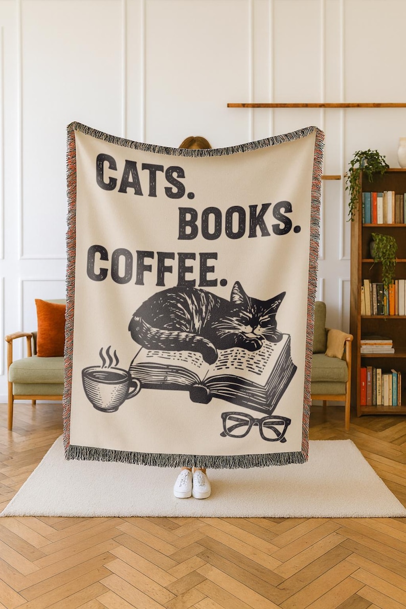 May include: Beige throw blanket with black text that reads "CATS. BOOKS. COFFEE." A black and white illustration of a cat sleeping on a book, a coffee cup, and glasses are also featured. The blanket has a fringed edge.
