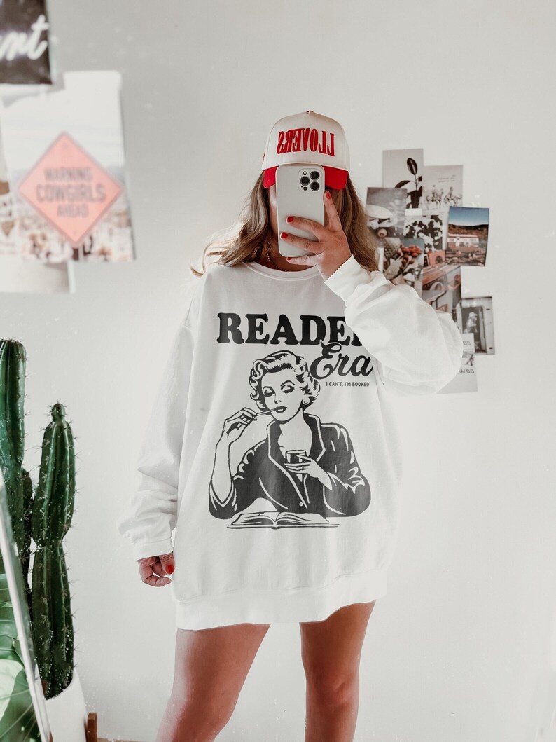 May include: White sweatshirt with a black and white illustration of a woman reading a book and drinking from a glass. The text "Reader Era" is printed above the illustration and the text "I can't, I'm booked" is printed below the illustration.