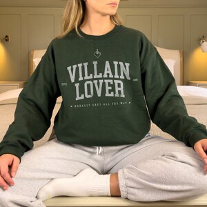 May include: A dark green sweatshirt with the text "VILLAIN LOVER" in white lettering. The sweatshirt has a small heart with a dagger through it above the text. The text "ESTD 2008" is below the text "VILLAIN LOVER". The text "MORALLY GREY ALL THE WAY" is below the text "ESTD 2008".