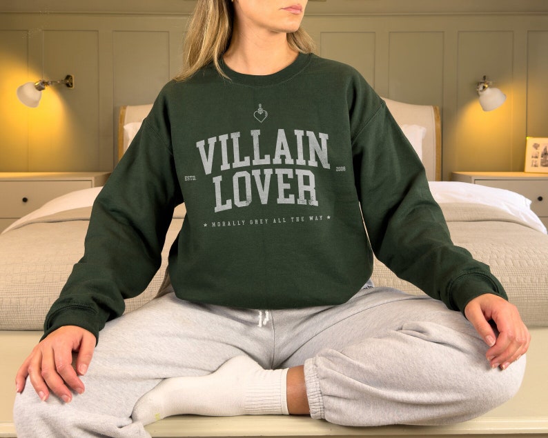 May include: A dark green sweatshirt with the text "VILLAIN LOVER" in white lettering. The sweatshirt has a small heart with a dagger through it above the text. The text "ESTD 2008" is below the text "VILLAIN LOVER". The text "MORALLY GREY ALL THE WAY" is below the text "ESTD 2008".