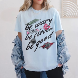Puede incluir: Camiseta azul claro con un estampado gr&aacute;fico de cartas con el texto "be wary be clever be good".