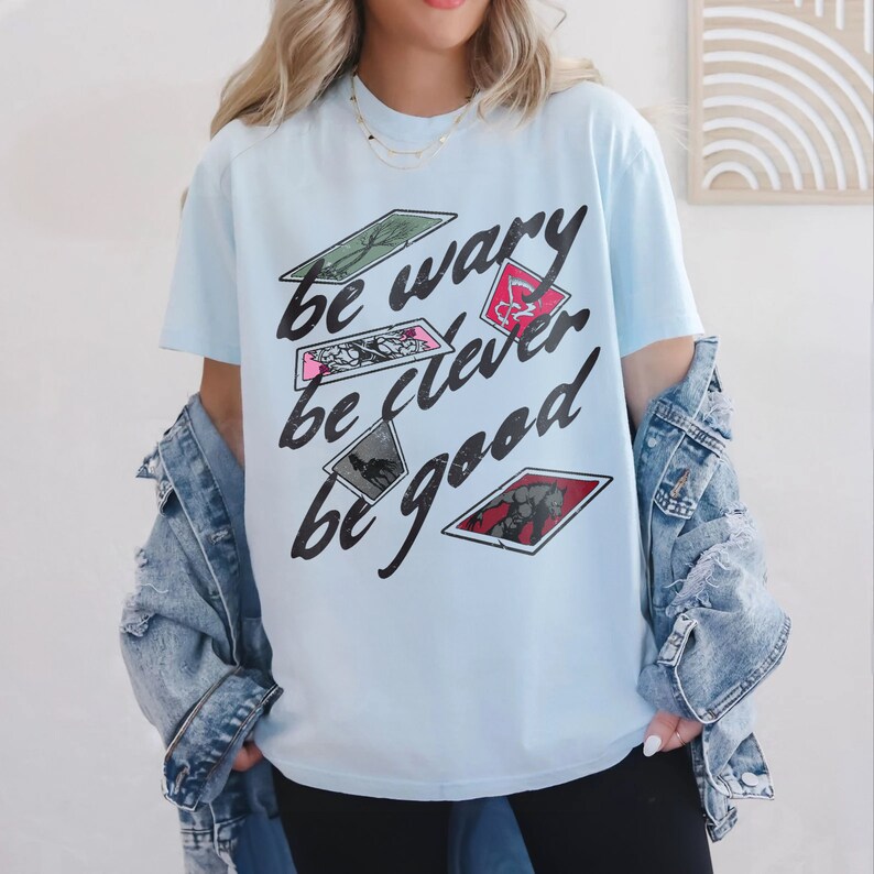 Puede incluir: Camiseta azul claro con un estampado gr&aacute;fico de cartas con el texto "be wary be clever be good".